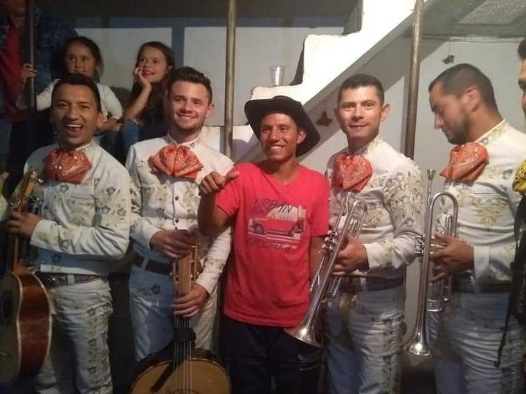 Mariachi juvenil con trompetas en una serenata a domicilio, junto a un joven de camiseta roja que levanta el pulgar y niñas mirando desde la escalera